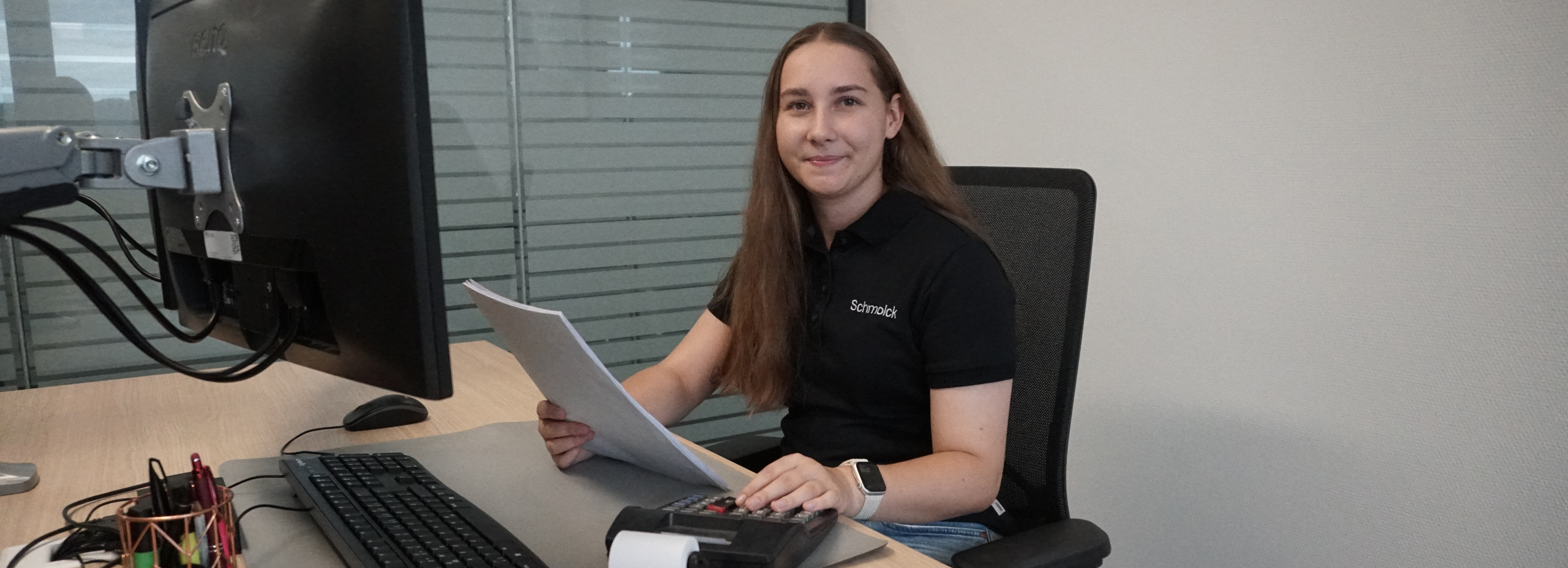 Auf dem Bild ist unsere Auszubildenede Laura Maier zusehen, die den Beruf als Bürokauffrau präsentiert. Sie sitzt an einem Schreibtisch, tippt was ein und hält ein Blatt in der Hand