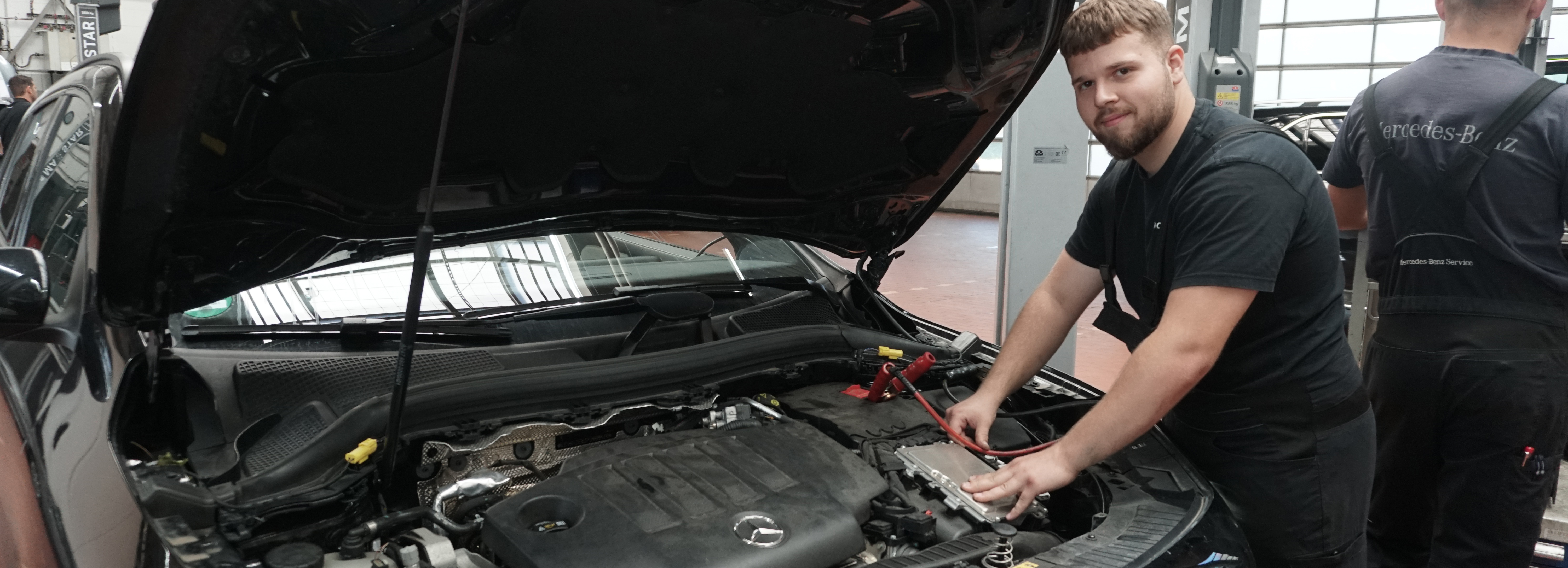 Auf dem Bild ist ein Auszubildender in der Merzedes Werkstatt zusehen, der die Batterie im Motorraum läd